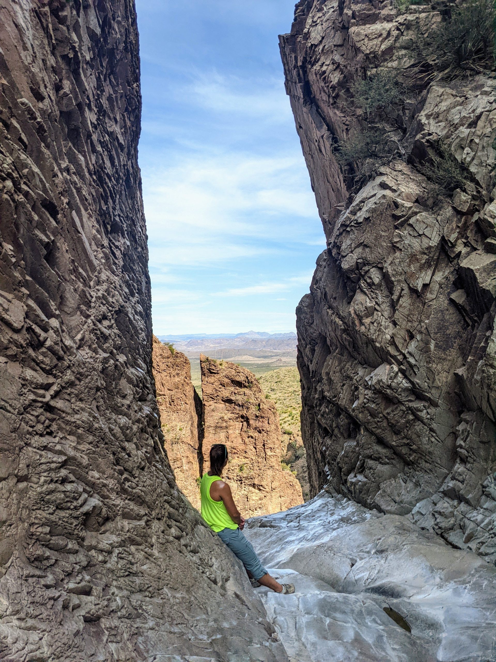 A woman rests against the wall of a narrow slot canyon, looking out from the top of the dry waterfall onto the vast desert below.