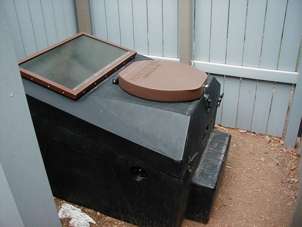 A green and brown composting toilet with a small step, partially enclosed by a fence.